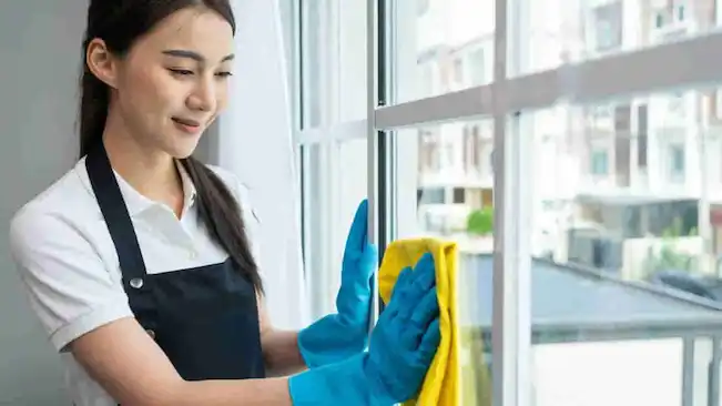 Professional house cleaning services in Singapore – a female cleaner wearing an apron and gloves carefully wiping a clean window frame.